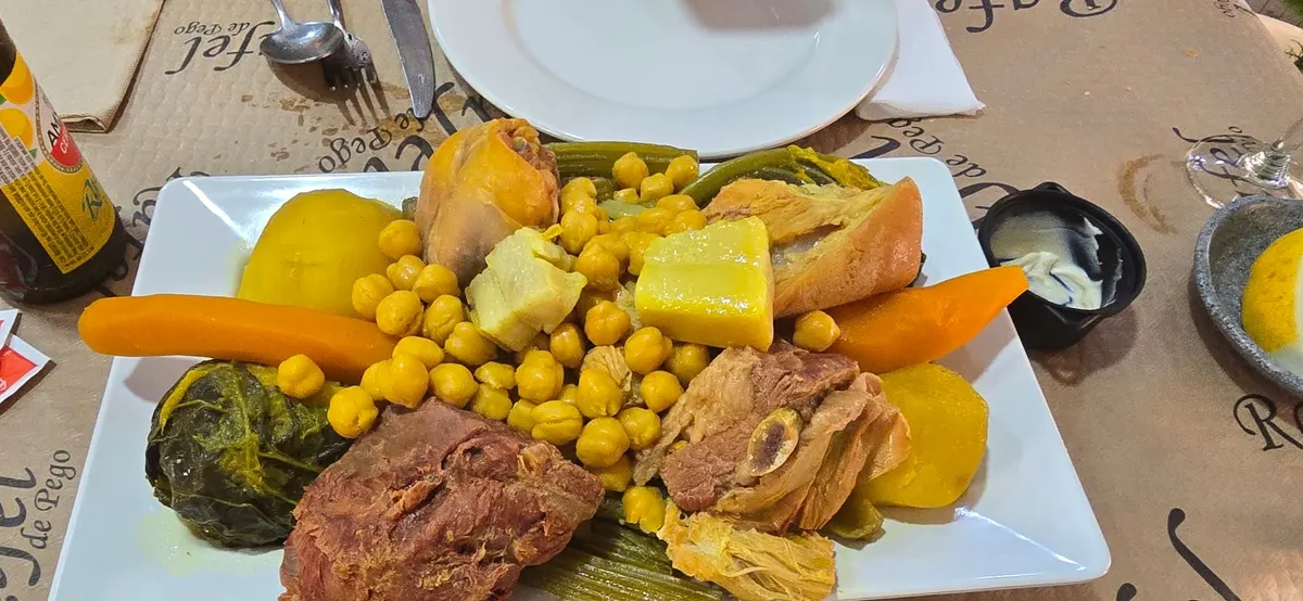 Fuente cuadrada de puchero con garbanzos, verduras y carnes sobre un mantel de papel con el nombre de Rafel